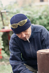 Close-up portrait of a man in a beanie and yellow glasses, smoking a cigarette outdoors. Smoke rises as he looks down, depicting a habit or addiction in a casual outdoor setting.