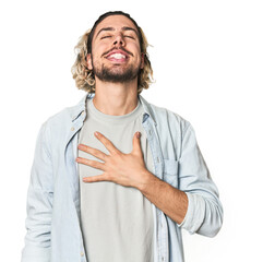 Young Caucasian man in studio setting laughs out loudly keeping hand on chest.
