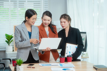 Three Asian women in formal suits collaborate at a desk, discussing documents in a modern office....