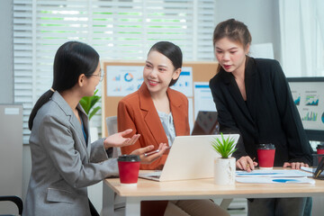 Three Asian women in formal suits collaborate at a desk, discussing documents in a modern office....