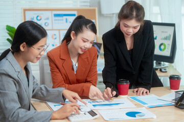 Three Asian women in formal suits collaborate at a desk, discussing documents in a modern office. teamwork, strategy planning, creative problem-solving in professional business environment.