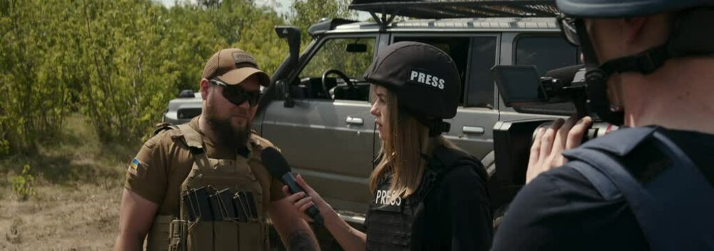 Female journalist wearing protective vest and helmet interviews Ukrainian soldier at frontline. Concept of war journalism, information gathering, work in conflict zone, safety, Ukraine.