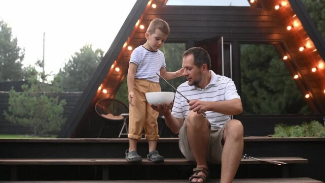 dad and little son cooking shashlik, barbecue, grilled meat in the yard of the house in the summer, threading meat on skewers against the background of a country house in nature in the summer