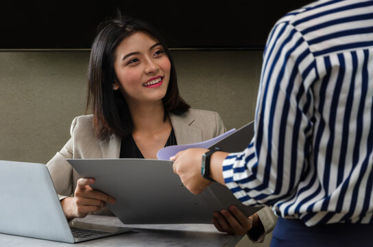 Asian female in beige blazer smiles warmly while receiving file folder with documents from colleague in striped shirt and sitting at office desk with laptop