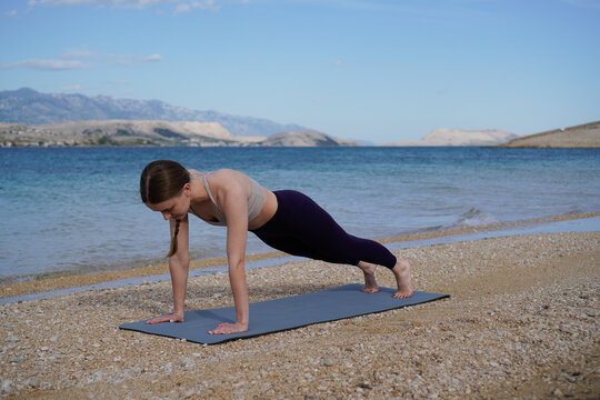 Fit woman holding a high plank pose during yoga practice on a coastal beach, showing strength and determination. - Powered by Adobe