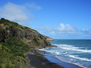 Fototapeta premium At Muriwai Beach, New Zealand, a rugged, greenery-covered rocky cliff crowned with small buildings overlooks a dark sandy shore where waves crash gently and beachgoers stroll beneath a clear sky speck