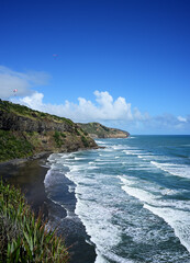 At Muriwai Beach, New Zealand, lush green rocky cliffs overlook a dark sandy shore where crashing, foamy waves animate a clear, cloud-dotted sky with two paragliders soaring overhead.