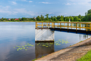 A bright wooden pier stretches over the calm lake in Trzebionka, Poland, offering a peaceful...