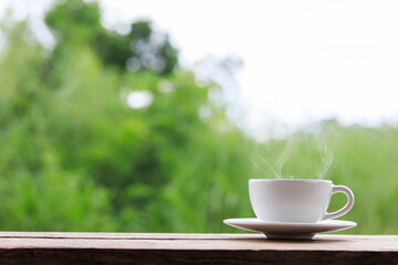 Hot coffee cup, on wooden table, in garden, green leaves blurred as background, white coffee cup