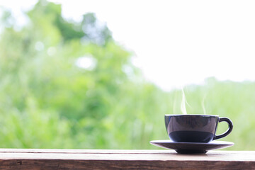 Hot coffee cup on wooden table in garden, green leaves blurred as background, black coffee cup