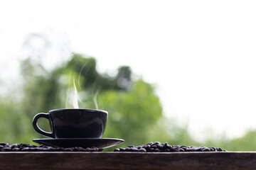 Hot coffee cup and coffee beans on wooden table in garden. Green leaves blurred as background. Black coffee cup.