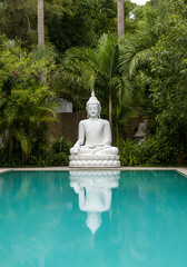 white Buddha statue sitting peacefully by a turquoise pool, surrounded by lush tropical foliage and palm trees in a serene garden. 