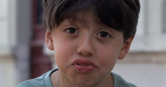 Playful young boy making funny face with tongue out and string of saliva hanging from mouth, looking directly at camera with wide eyes and silly expression outdoors