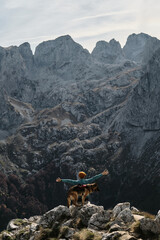A female traveler raises her arms in triumph while standing with her German Shepherd atop a rocky...