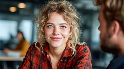 Fototapeta premium A young woman with curly hair and a warm smile looks confidently into the camera while seated in a modern office environment, surrounded by a professional vibe and soft lighting.