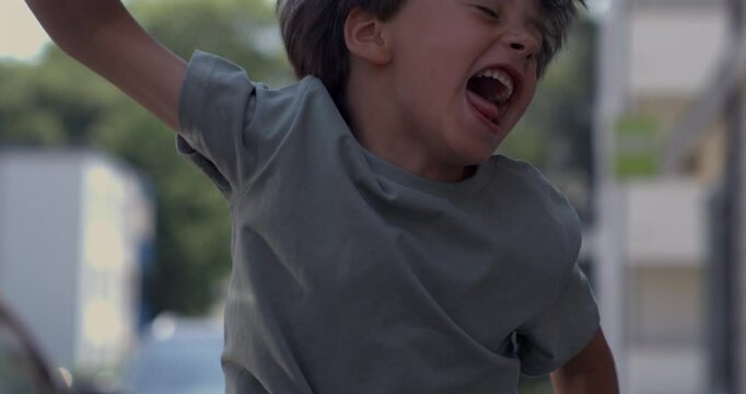 Energetic young boy mid-jump with fists clenched and mouth open in loud joyful scream, captured in super slow motion on street in broad daylight