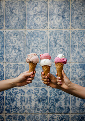 three hands holding artisanal gelato cones with dual pastel flavors in front of a vintage blue-and-white tiled wall. 