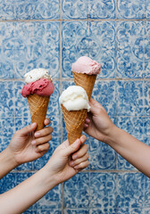 three hands holding artisanal gelato cones with dual pastel flavors in front of a vintage blue-and-white tiled wall. 