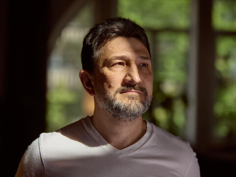 Emotional depth. Caucasian man in a white shirt gazes thoughtfully through a sunlit window in a cozy room. Concept of introspection and contemplation in a peaceful setting.