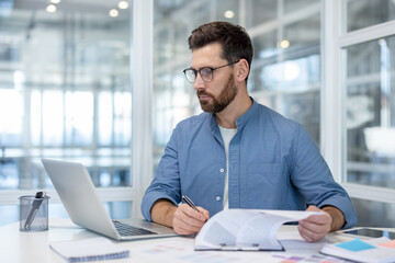 A professional man, seated at a desk, engrossed in paperwork, laptop, and working in office.