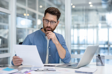 A man in a blue shirt and glasses reviews documents, thoughtfully considering information.