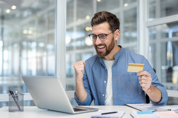 A man celebrates a successful online purchase, holding a credit card near a laptop in an office...