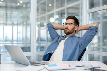 A relaxed businessman in a modern office takes a moment to unwind, resting with his hands behind his head near a laptop.