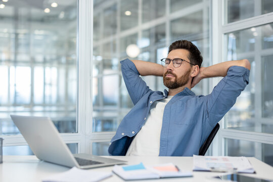 A relaxed businessman in a modern office takes a moment to unwind, resting with his hands behind his head near a laptop. - Powered by Adobe