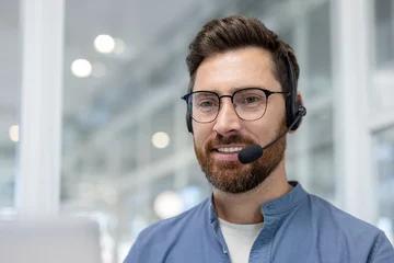 Fotobehang A man with a headset and glasses smiles, likely assisting customers, set against a bright, blurred background. © Liubomir