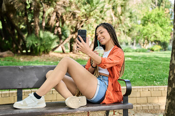 Naklejka premium Young latina woman taking a selfie while enjoying a refreshing drink in the park
