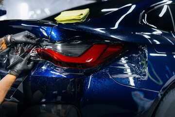 Installing PPF tint protective film on the rear light of a blue car during a professional detailing process in a modern, well-lit car repair shop. Marketing, advertising or press articles.
