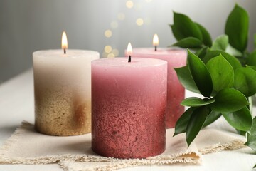 Burning candles and green leaves on white table against blurred lights, closeup