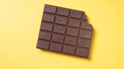 A bitten dark chocolate bar with neatly divided squares placed on a bright yellow background,...
