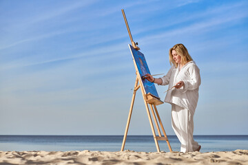 Young woman artist stands near ocean, painting on easel. Serene beach environment complements her...
