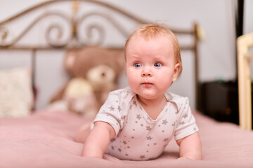Fototapeta premium Young female child with light hair, playful expression, star-patterned pajamas. Pink bedding, blurry teddy backdrop, soft lighting, cozy home setting, warm atmosphere