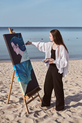 Young female painter works on canvas by ocean, capturing seascape inspiration. Her focused...