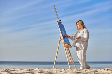 Young woman artist stands near ocean, painting on easel. Serene beach environment complements her...