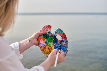 Young female artist blends vibrant paint on palette by serene seaside. Calming ambiance and natural...