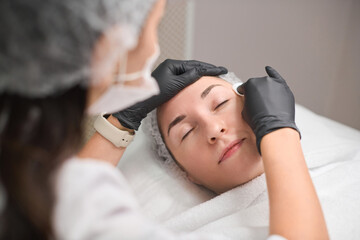 Young adult woman receives skincare treatment, lying comfortably with closed eyes. Female beautician wipes client skin of face with cotton pad. Serene clinic environment and professional care