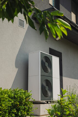Vertical outdoor heat pump unit installed next to a house wall, surrounded by green shrubs in a...