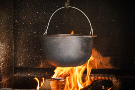 Iron cauldron hanging over burning firewood inside rustic fireplace
