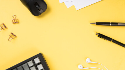 Flat lay of office essentials including a black mouse, keyboard, pens, paper clips, earphones, and white paper sheets arranged neatly on a vibrant yellow background