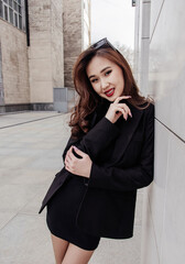 A smiling woman with braces and long hair poses in a black blazer outdoors by a modern building