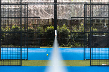 A padel court with blue and green turf is being cleaned by an automated sprayer positioned at the...