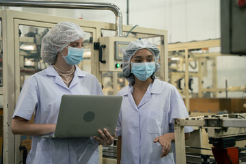 Female engineers in medical mask collaborate. Laptop use. Machinery inspection. Cleanroom environment. Teamwork evident, Two workers review data, discussing project