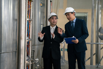 Businessmen in hard hats examine industrial machinery. Large metal tanks stand nearby....