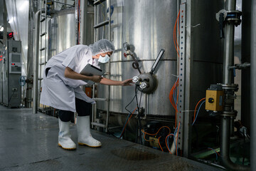 Female worker in protective uniform inspects machinery. Stainless steel tanks fill industrial...