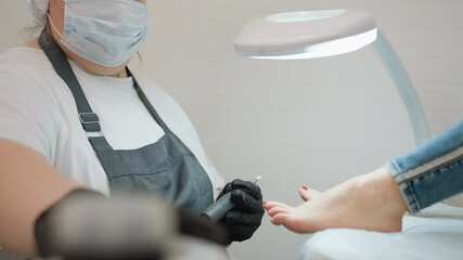 Close up of nail technician wearing mask and gloves connecting electric filer tool while client rests foot with red polish on padded surface under bright lamp in clean professional salon environment