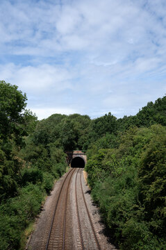 A railway track leading into a dark tunnel surrounded by lush greenery, with a bright summer sky above.