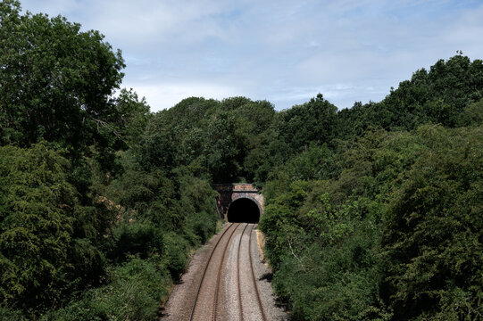 A railway track leading into a dark tunnel surrounded by lush greenery, with a bright summer sky above.